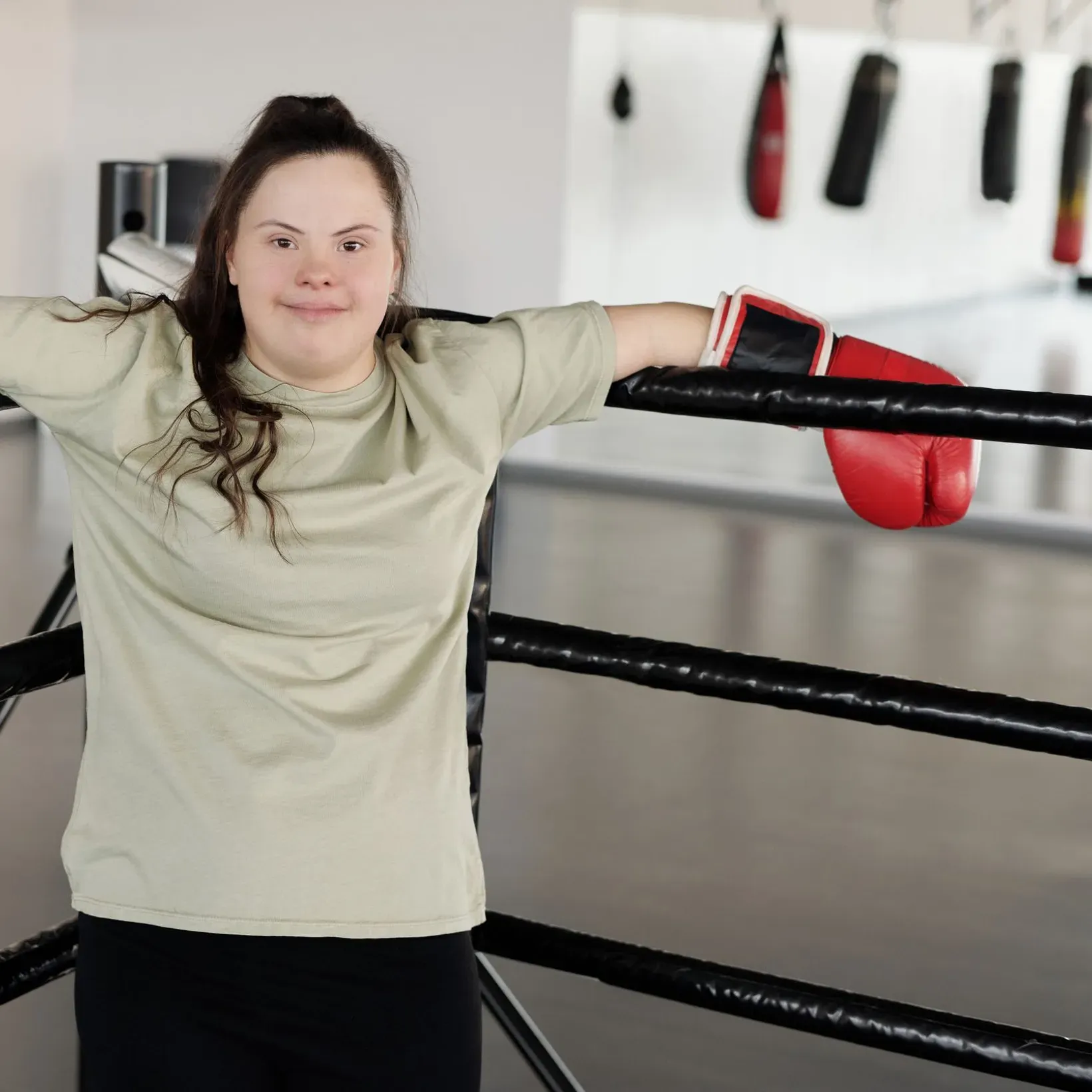 A young woman with Down syndrome leaning on the ropes of a boxing ring in a gym