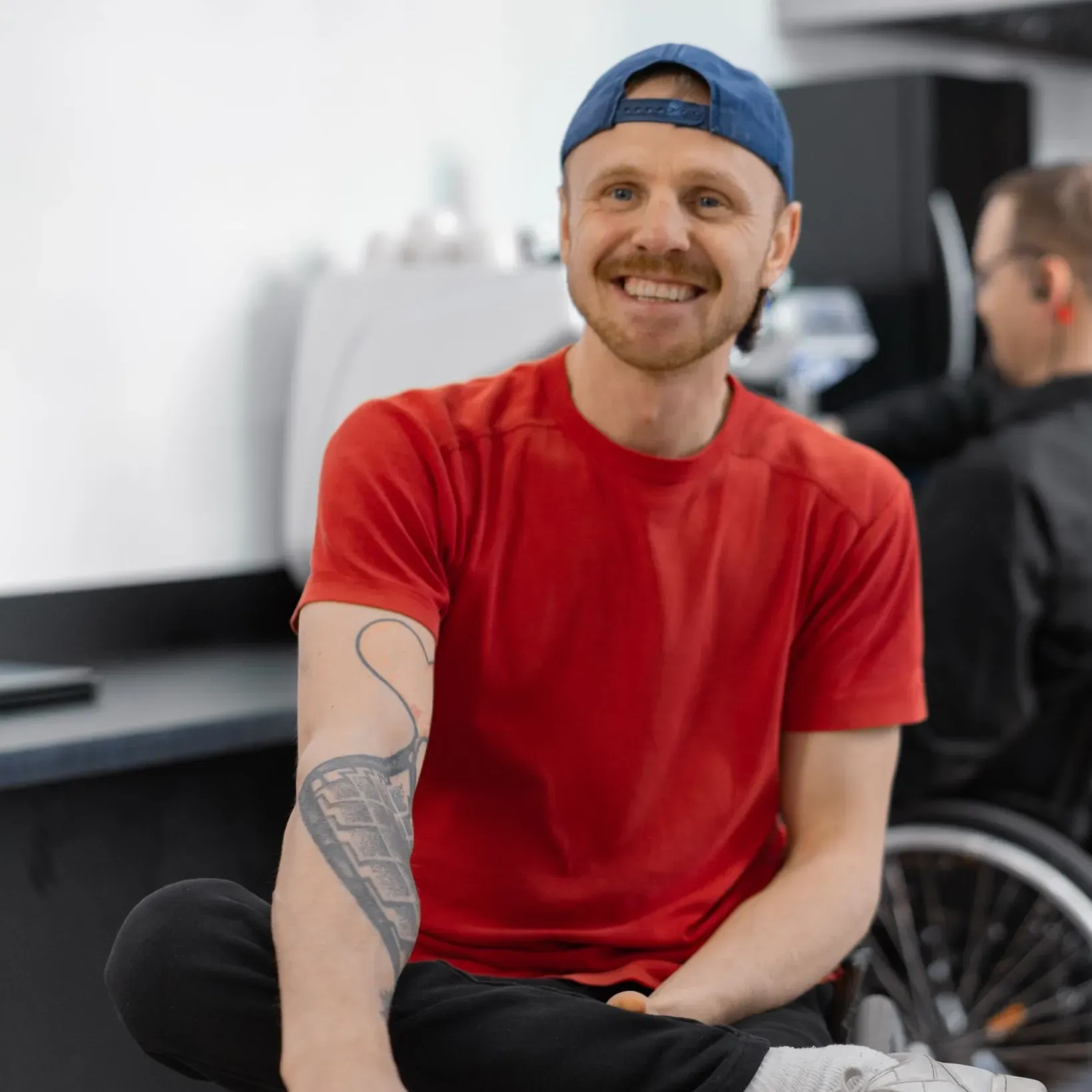 A barista smiling in a cafe, a colleague in a wheelchair working behind him